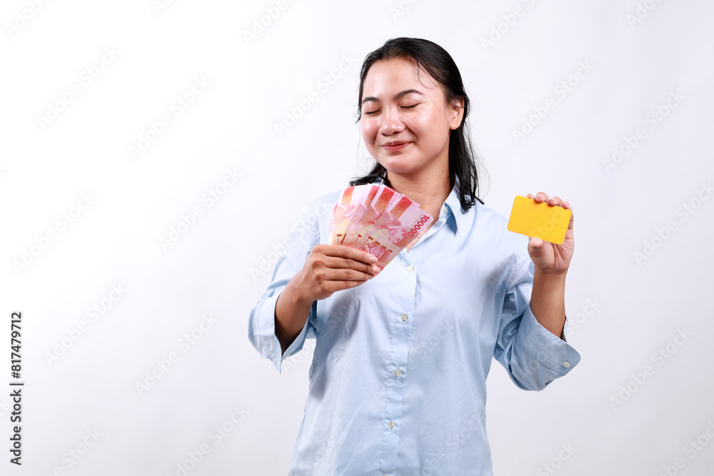 Happy asian woman holding credit card and Indonesian money rupiah, smiling, posing against white studio background Happy asian woman holding credit card and Indonesian money rupiah, smiling, posing against white studio background
