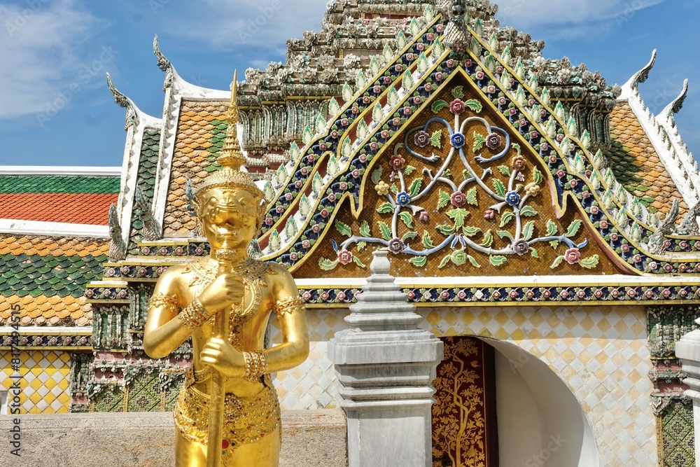 Obraz premium Gold chimera statue in front of a decorative side gate between the Temple of the Emerald Buddha (Wat Phra Kaew) and the Grand Palace in Phra Nakhon, Bangkok, Thailand on a fine day with blue skies