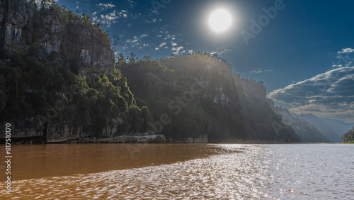 A red-brown exotic river with steep banks. Green vegetation on the sheer limestone slopes. The sun is shining in the blue sky. Picturesque clouds. Ripples and highlights on calm water. Madagascar.  photo