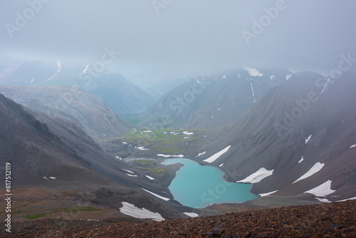Most beautiful turquoise alpine lake in stony rocky green valley against mountain range silhouette in rainy low clouds in gray sky. Large snowy mountains in light transparent mist in rain bad weather.