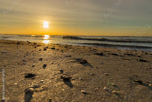 Ground level shot of sunrise over sandy beach with focus on sand, pebbles and shells in foreground and golden sun rising in a hazy blue sky with reflections on sea.