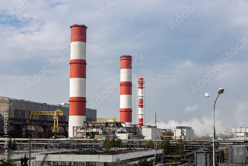 Red and white chimneys of a power plant with emissions, pipes, buildings and blue sky; chimneys of a fossil fuel burning power station with visible pollution.