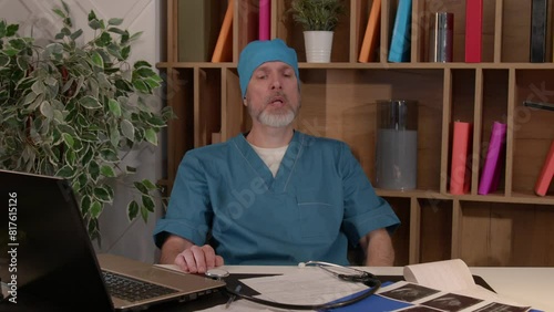 Exhausted handsome middle aged male surgeon in uniform taking a break and relaxing at desk in medical office after hard surgery, being tired and overworked