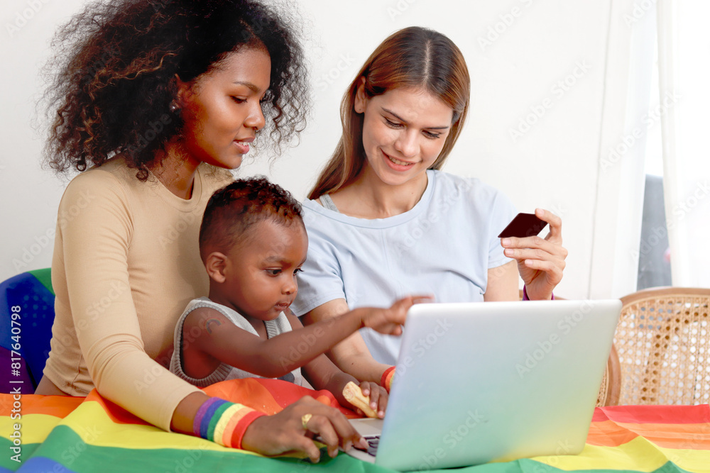 Happy LGBT family, homosexual lesbian couple with rainbow flag wristband and adopted child use laptop computer together. Beautiful woman and African girlfriend search for pride celebration gifts. Happy LGBT family, homosexual lesbian couple with rainbow flag wristband and adopted child use laptop computer together. Beautiful woman and African girlfriend search for pride celebration gifts.