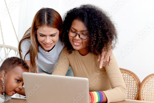 Happy LGBT family, homosexual lesbian couple with rainbow flag wristband and adopted child use laptop computer together. Beautiful woman and African girlfriend search for pride celebration gifts.