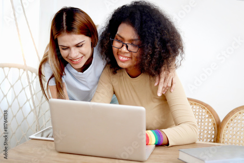 Happy homosexual lesbian couple with rainbow flag wristband using laptop computer together. Beautiful woman and African girlfriend search for pride celebration gifts online. Romantic LGBT lover family