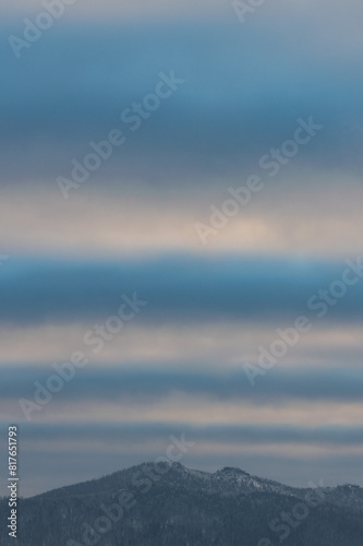 Unusual clouds pass in wide stripes across the blue sky, illuminated by sunlight above the top of a mountain overgrown with forest