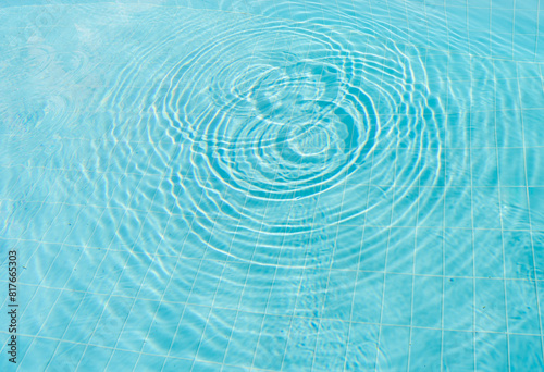 Clear blue swimming pool water with ripples radiating outward, creating mesmerizing pattern on the tiled pool floor. Image captures the tranquil and refreshing essence of calm, sunny day by the pool.