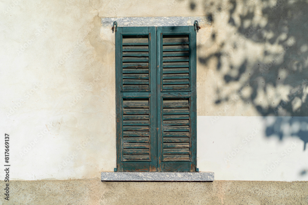 Fototapeta premium old teal shutters on a weathered window set against a textured white wall with dappled shadows