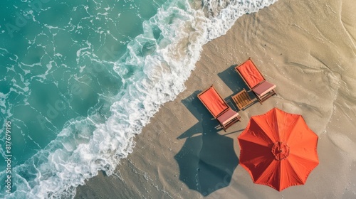 A tranquil scene from above displays two sun loungers and a vibrant red umbrella on a sandy beach lapped by gentle waves