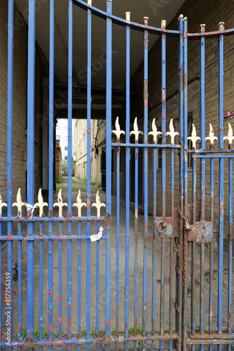 Metal gate in blue with yellow finials. The gate is padlocked with a chain, and several areas are now rusty or flaking paint revealing red underneath.