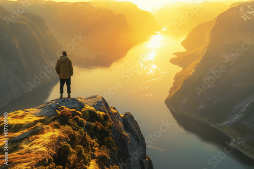 person standing on the edge of an ancient mountain  overlooking the vast fjords in golden sunlight at sunrise. The individual is dressed casually with their back to camera as they