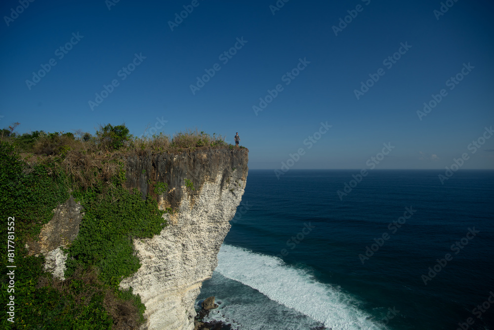 A person stands atop a towering seaside cliff overlooking the ocean under a clear blue sky. A person stands atop a towering seaside cliff overlooking the ocean under a clear blue sky.