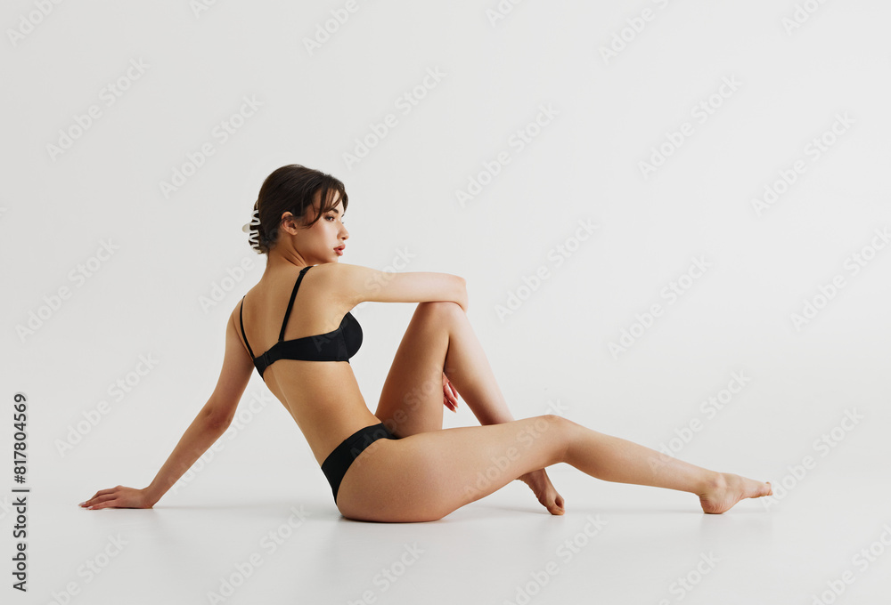 Elegant youth. Beautiful young girl with slim, fit, perfect body shape sitting on floor in black underwear isolated on grey background. Concept of natural beauty, body care, sport, wellness, health