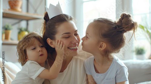 joyful mother being kissed by her daughter, with her son nearby, all smiling and playing in a bright home, suggesting a loving and playful family environment