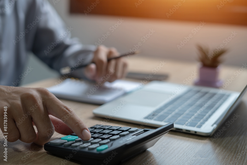 A person is using a calculator on a desk with a laptop and a potted plant. The person is focused on the calculator, possibly working on a math problem or a financial calculation A person is using a calculator on a desk with a laptop and a potted plant. The person is focused on the calculator, possibly working on a math problem or a financial calculation