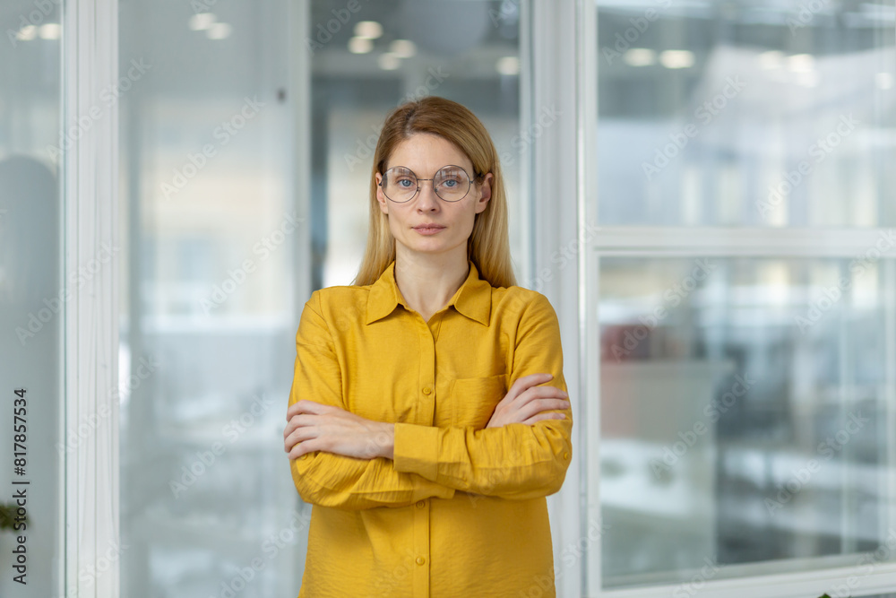 Confident businesswoman with glasses standing in modern office, arms crossed, looking at camera. Professional and determined appearance in contemporary workspace.