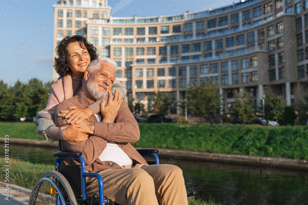 Fototapeta premium Old man in wheelchair walking with caregiver senior woman on road in park. Elderly family couple woman supporting embracing paralyzed man in chair for people with disability outdoor. Rehabilitation