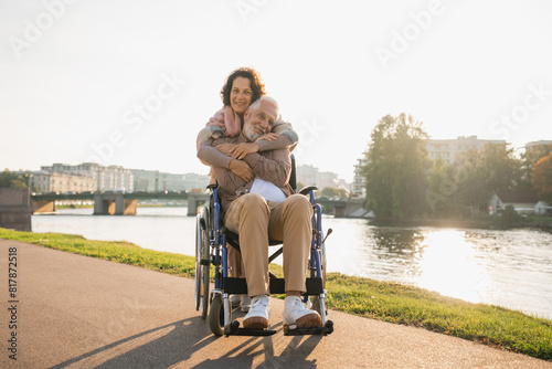 Old man in wheelchair walking with caregiver senior woman on road in park. Elderly family couple woman supporting embracing paralyzed man in chair for people with disability outdoor. Rehabilitation