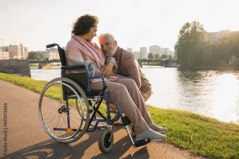 Senior woman in wheelchair walking with caregiver old man on road. Elderly family couple man supporting taking care of paralyzed woman in chair for people with disability outdoor. Rehabilitation Senior woman in wheelchair walking with caregiver old man on road. Elderly family couple man supporting taking care of paralyzed woman in chair for people with disability outdoor. Rehabilitation