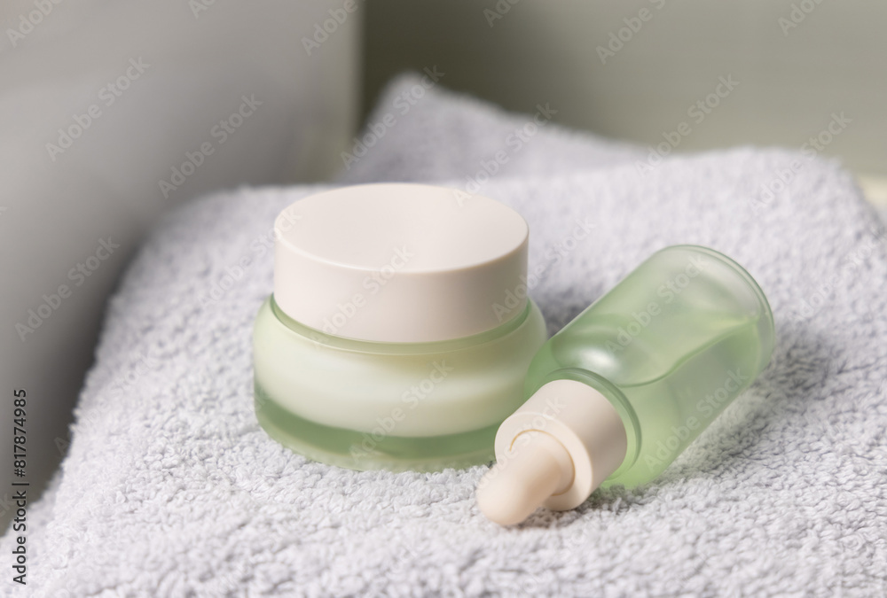 Light green dropper bottle and cream jar on light grey bath towel near basin closeup Light green dropper bottle and cream jar on light grey bath towel near basin closeup