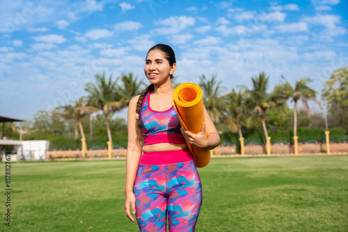 Fit young indian woman holding yoga mat in garden smiling. exercise, meditation, workout, outdoor, mental health, well being.