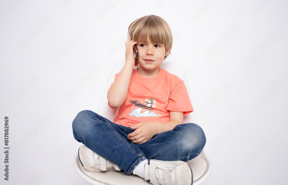 Little boy holding in hand a mobile cell phone sitting on chair isolated on white background. Little boy holding in hand a mobile cell phone sitting on chair isolated on white background.