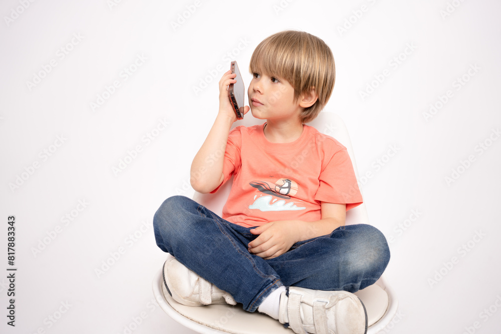 Little boy holding in hand a mobile cell phone sitting on chair isolated on white background. Little boy holding in hand a mobile cell phone sitting on chair isolated on white background.