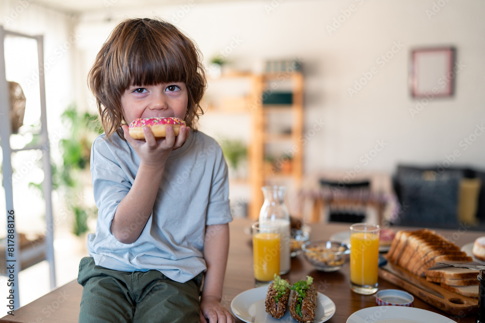 Portrait of a cute little boy eating donut during breakfast time at home. He is sitting on the table in the kitchen and looking at camera. Portrait of a cute little boy eating donut during breakfast time at home. He is sitting on the table in the kitchen and looking at camera.