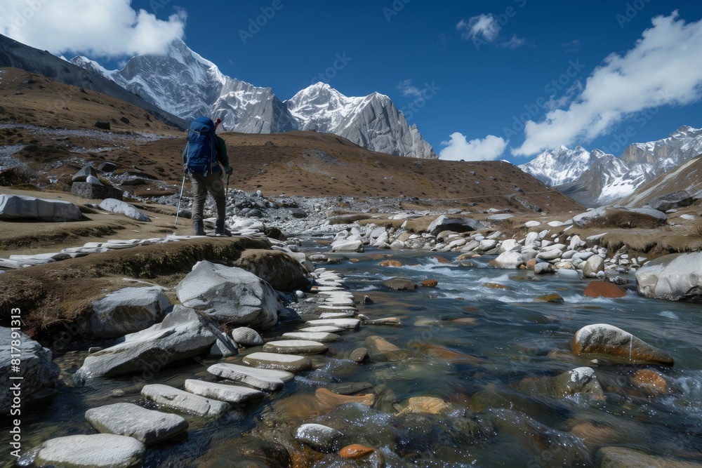 A trekker with a backpack and hiking poles crossing a mountain stream on a stone path, surrounded by snow-capped peaks and rugged terrain.