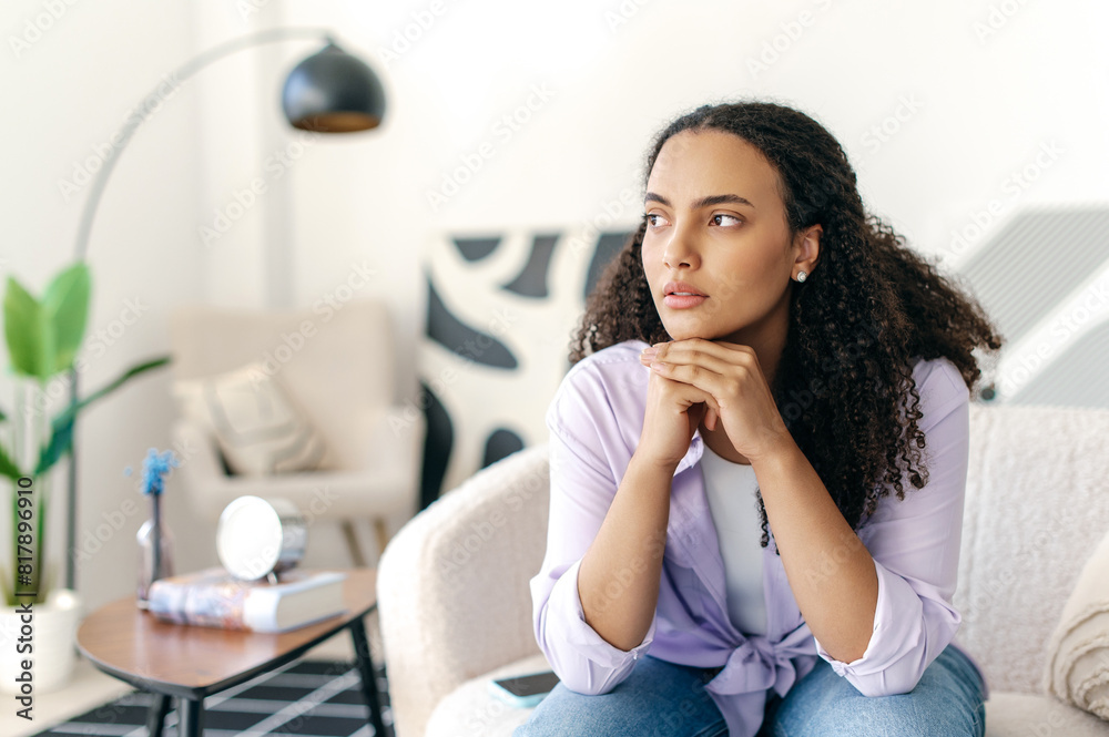 Worried depressed young brazilian or hispanic curly haired woman, sitting alone on a sofa, suffering from loneliness, feeling stressed, tired, thinks over bad news, problems in relationships Worried depressed young brazilian or hispanic curly haired woman, sitting alone on a sofa, suffering from loneliness, feeling stressed, tired, thinks over bad news, problems in relationships