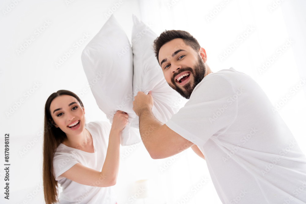 Obraz premium Photo of cheerful funky couple wear white t-shirts pillow fighting indoors apartment bedroom