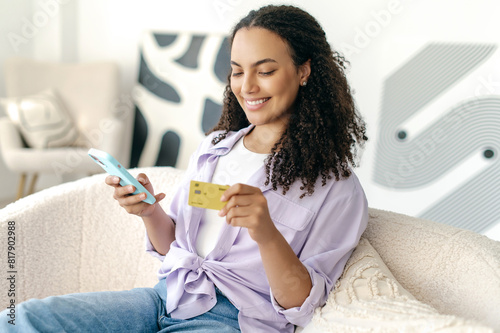 Online shopping concept. Positive attractive brazilian or hispanic curly haired woman using a credit card for shopping and banking online with a mobile phone while sitting at home on the couch, smiles