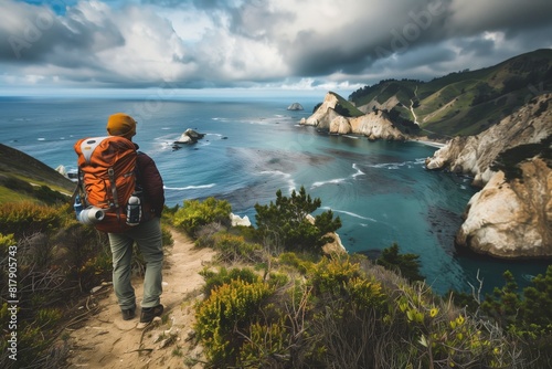 A hiker with an orange backpack stands on a coastal trail overlooking dramatic cliffs and a serene ocean under a cloudy sky.
