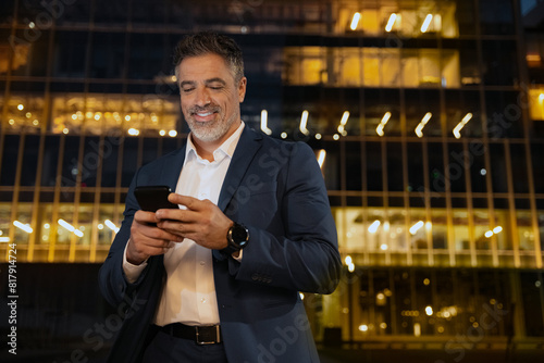 Smiling mature hispanic senior business man using app, holding smartphone cellphone at office centre at night. Indian or latin middle age male entrepreneur businessman working on mobile cell phone.
