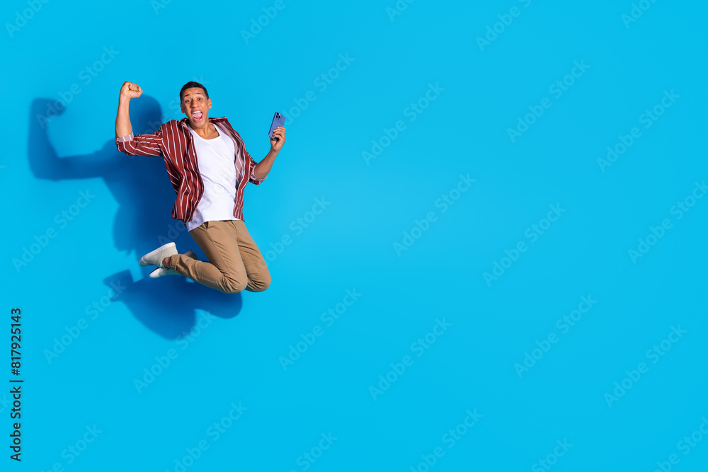 Full size photo of overjoyed man dressed striped shirt hold smartphone flying empty space win gambling isolated on blue color background