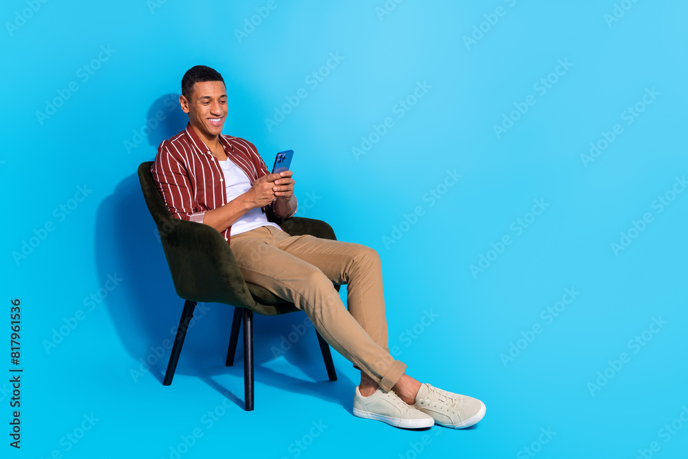 Full size photo of satisfied man dressed shirt over white t-shirt sit on armchair look at samrtphone isolated on blue color background Full size photo of satisfied man dressed shirt over white t-shirt sit on armchair look at samrtphone isolated on blue color background