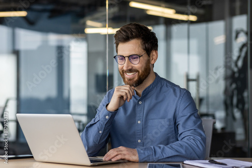 A smiling young man in glasses and a denim shirt is sitting in the office at a desk, working and talking on a video call online photo