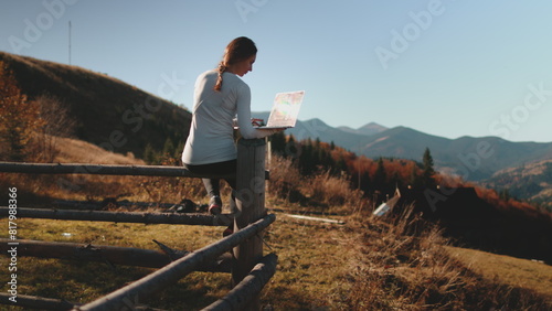 A woman stands confidently atop a hill  typing on a laptop computer. The wind ruffles her hair as she works against the backdrop of a stunning panoramic view.