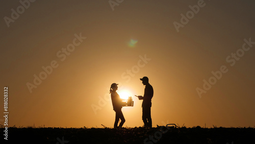 A woman with a box of seedlings and a man with a tablet stand in a field at sunset. Farmers discussing sowing season