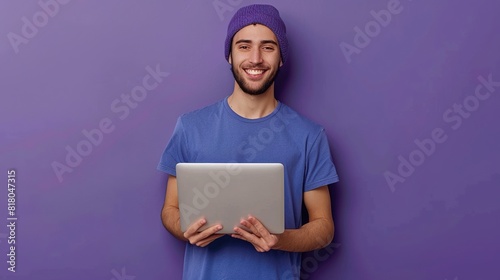 Young smiling businessman or student in blue t-shirt and beanie holding laptop while standing on violet background and looking at camera 