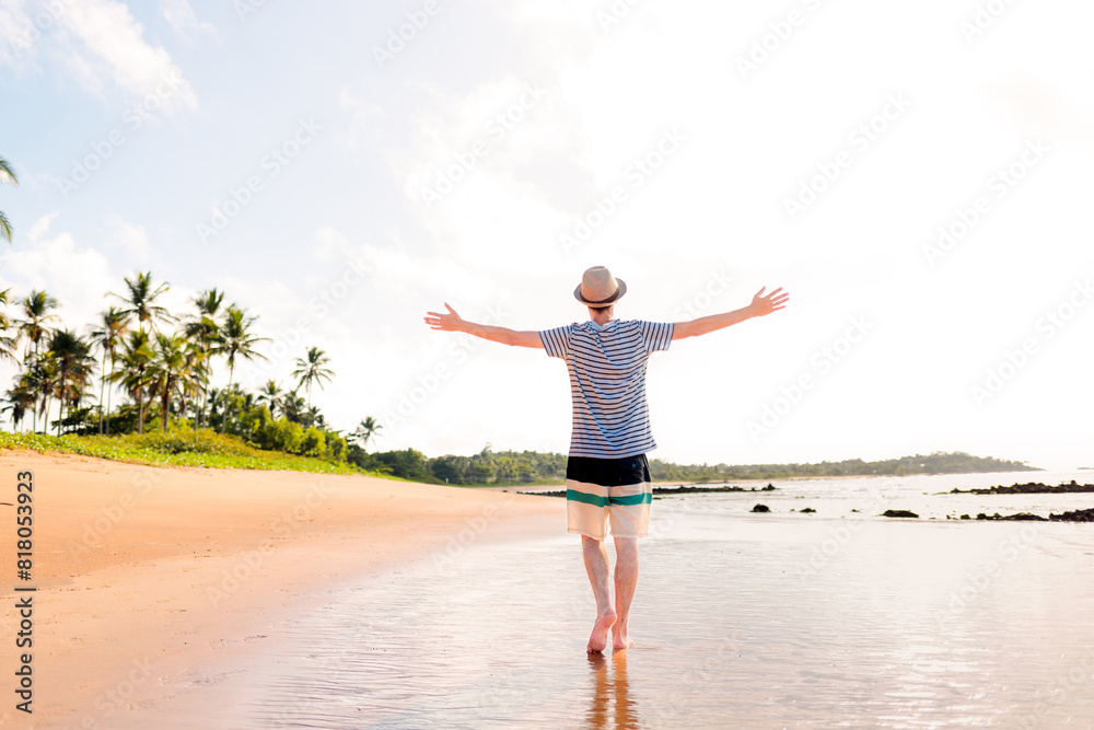 Brazilian man wearing sun hat, with his arms raised to express gratitude and happiness for enjoying summer vacation on a paradise beach Brazilian man wearing sun hat, with his arms raised to express gratitude and happiness for enjoying summer vacation on a paradise beach
