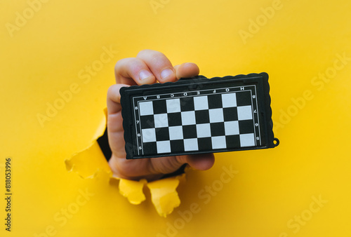 A man's hand emerges from a ragged hole in the yellow paper and holds a small chessboard. The concept of intellectual development and games that develop thinking.
