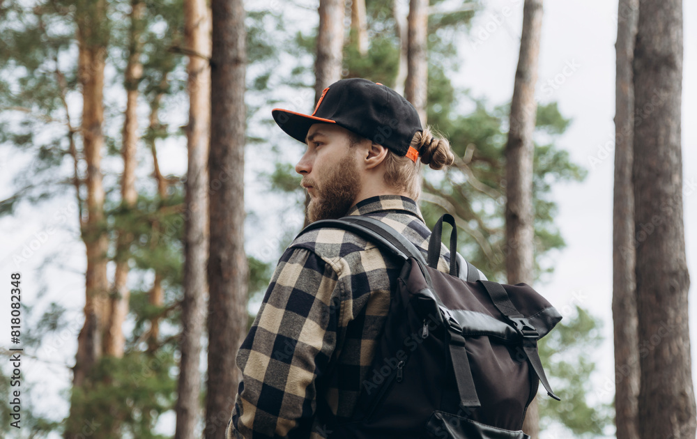 Bearded young man in a cap and with a backpack in a pine forest, hiking concept.