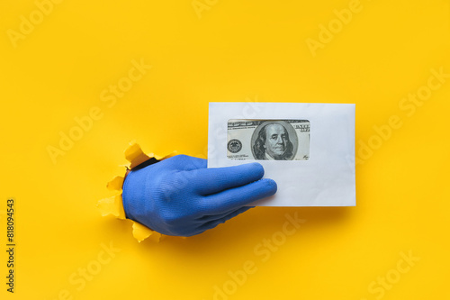 The right male hand in a blue fabric work glove holds an envelope with dollar bills (money). Torn hole in yellow paper. Concept of guest worker, handyman, gray illegal salary for work. Copy space. photo