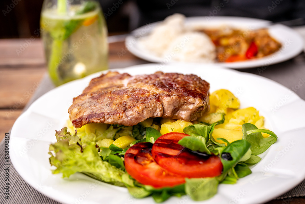 Pork steak on potato salad with vegetable on first white plate, drink and second meal on table. Pork steak on potato salad with vegetable on first white plate, drink and second meal on table.
