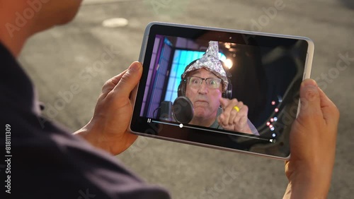A man, holding a tablet PC outside, watches a conspiracy theorist podcast.  	