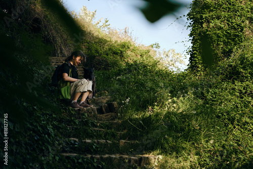 Young woman with black mix breed dog sits and poses on an old stone staircase in the middle of a green spring park. A dog with female owner rests outside on a summer sunny day in shadows of trees.