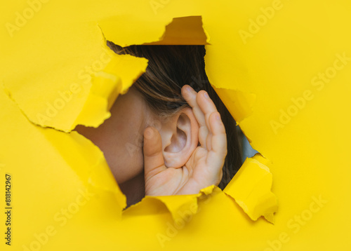 Female left ear and hand close-up. Copy space. Torn paper, yellow background. The concept of eavesdropping, espionage, gossip and the yellow press.