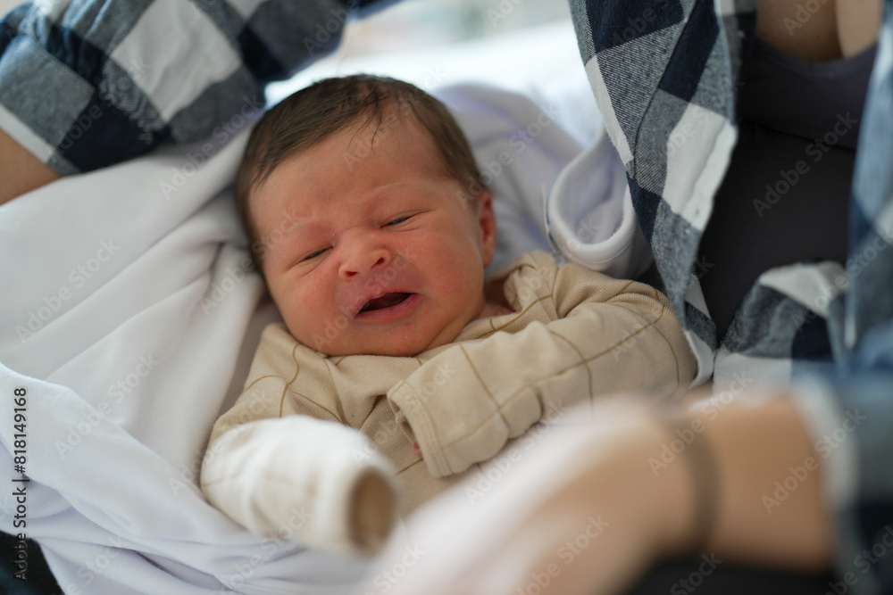 Baby dressed in a beige outfit, crying in mother's arms while lying on a bed. The mother, wearing a plaid shirt, looks down with concern and care, highlighting a tender moment of parenting